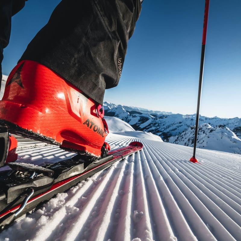 Nahaufnahme eines roten Skischuhs in der Bindung auf frisch präparierter Piste mit Bergblick